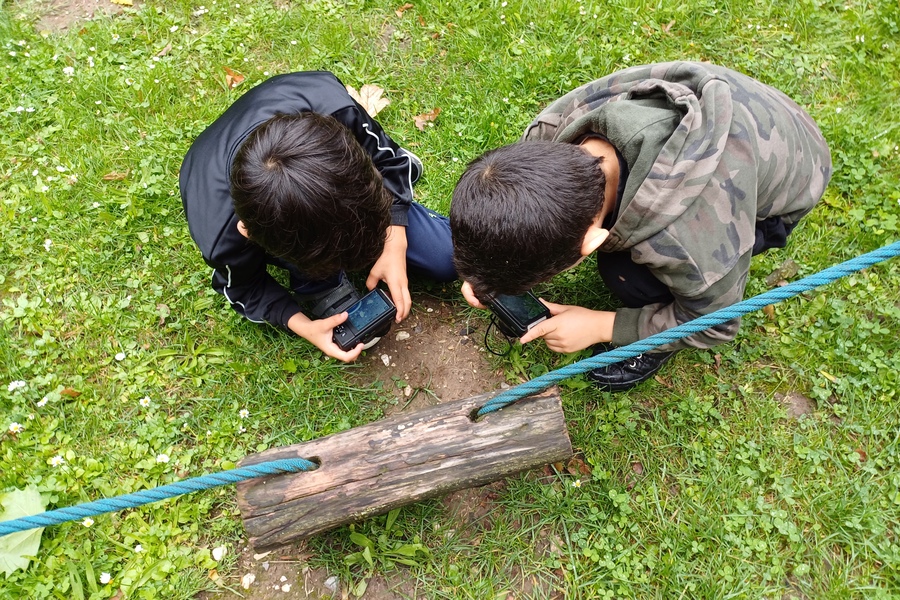 Zwei Jungen hocken auf dem Boden im Gras. In der Hand halten beide Kameras, mit denen beide Fotos machen.