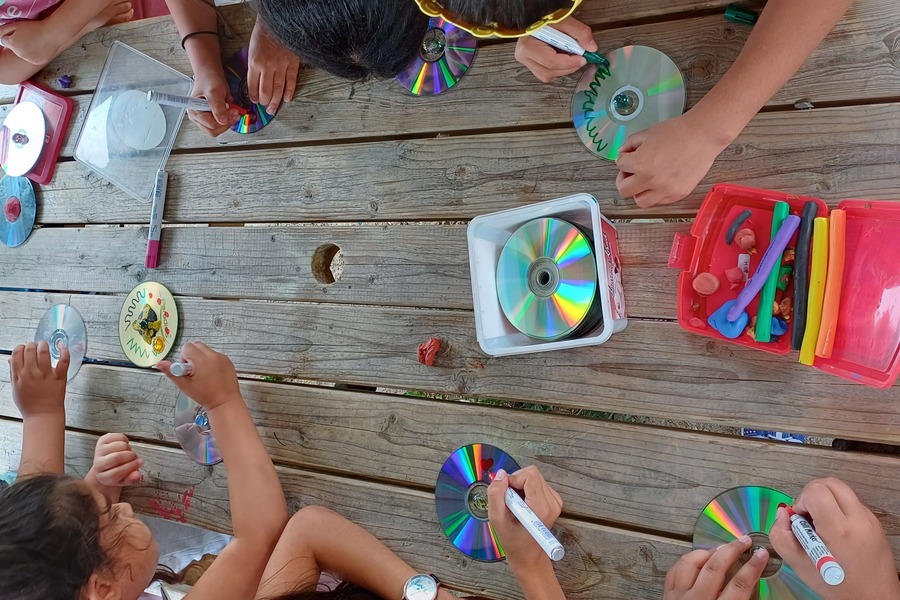 Tisch in der Mitte mit Kindern auf beiden Seiten des Tisches. Die Kinder malen mit Stiften auf CDs.