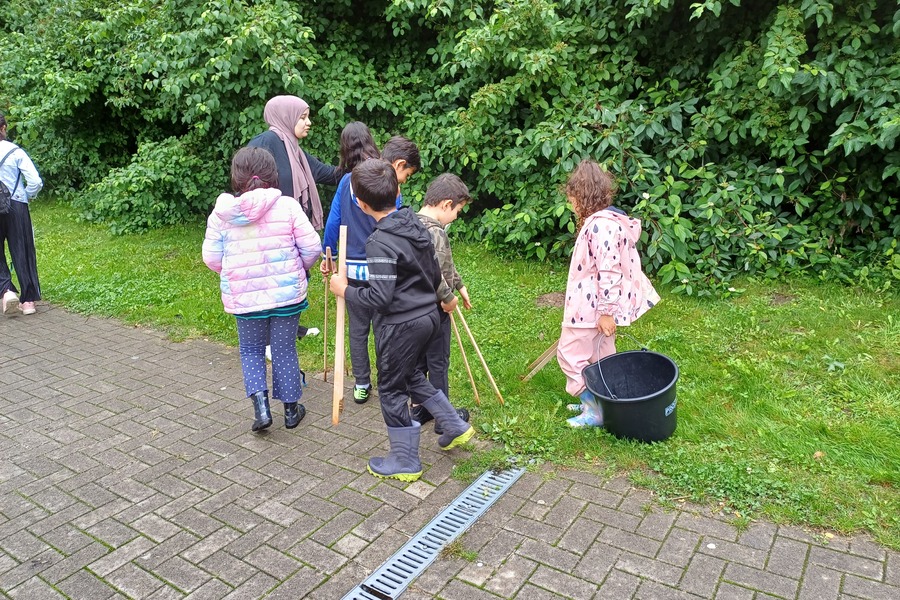 Kinder laufen mit einer Erzieherin auf einem gepflasterten Weg. An der Seite des Wegs sind Gras und Büsche. Die Kinder halten EImer und Müllzangen aus Holz in der Hand.