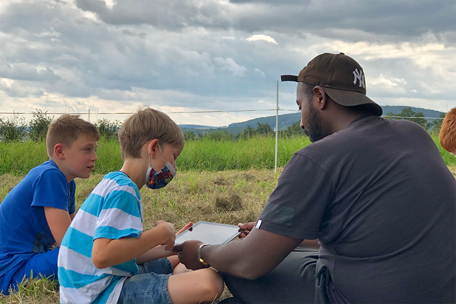 Betreuer und zwei Jungen sitzen auf einem Feld und schauen auf ein Tablet.